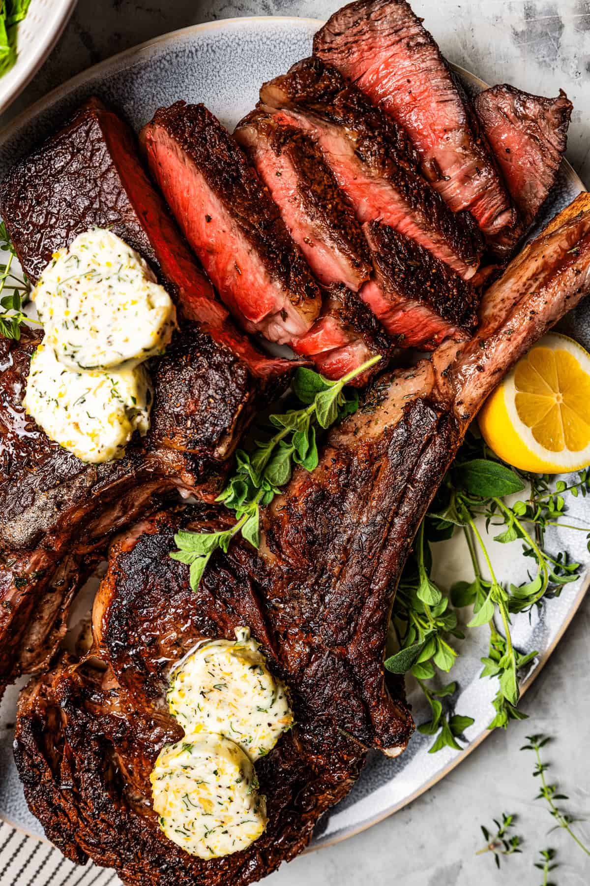 Overhead view of sliced tomahawk steak topped with compound butter on a platter, garnished with fresh herbs and lemon slices.