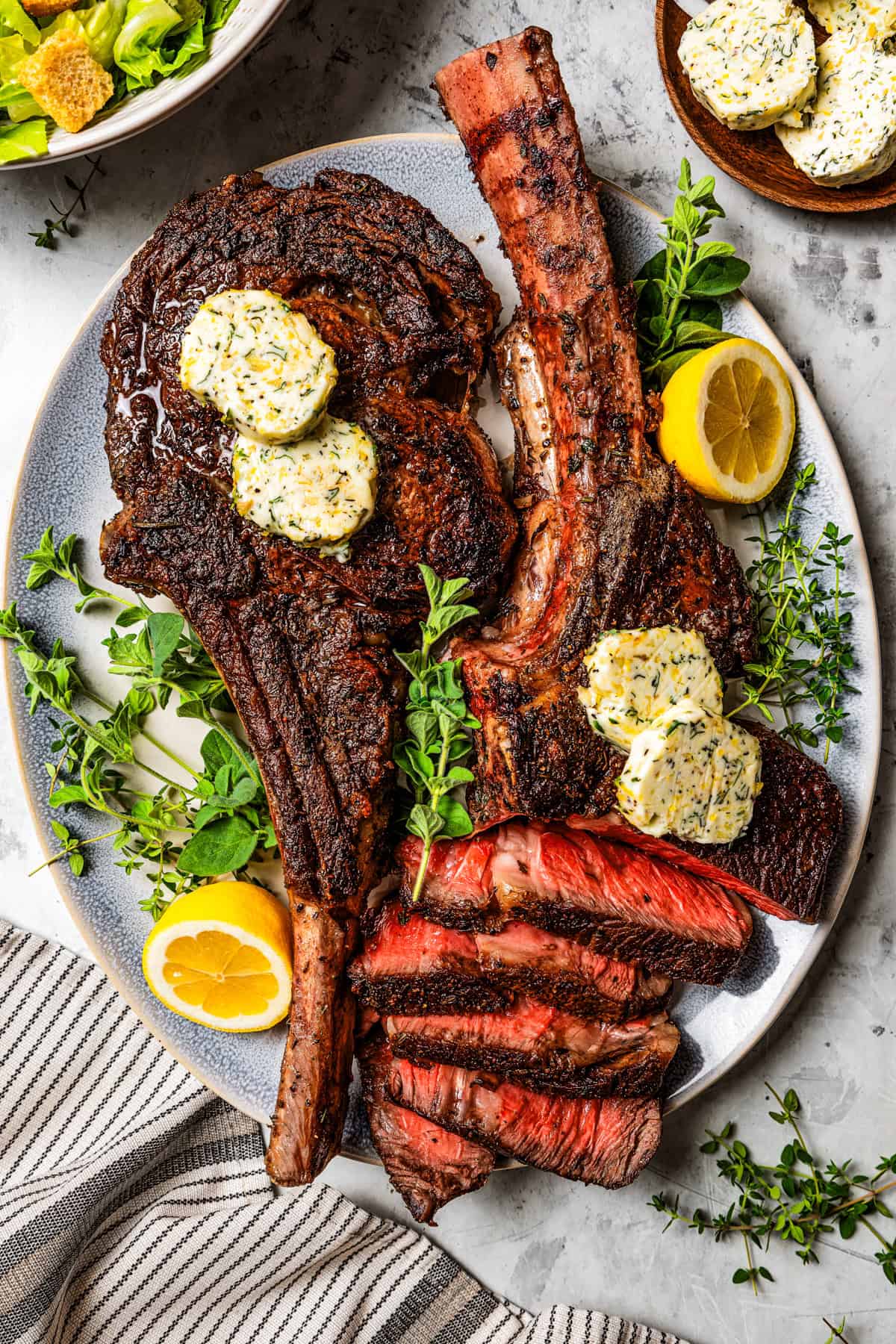 Two tomahawk steaks served on a platter, with one of the steaks already cut into slices, and pats of herbed butter resting on top of both steaks.