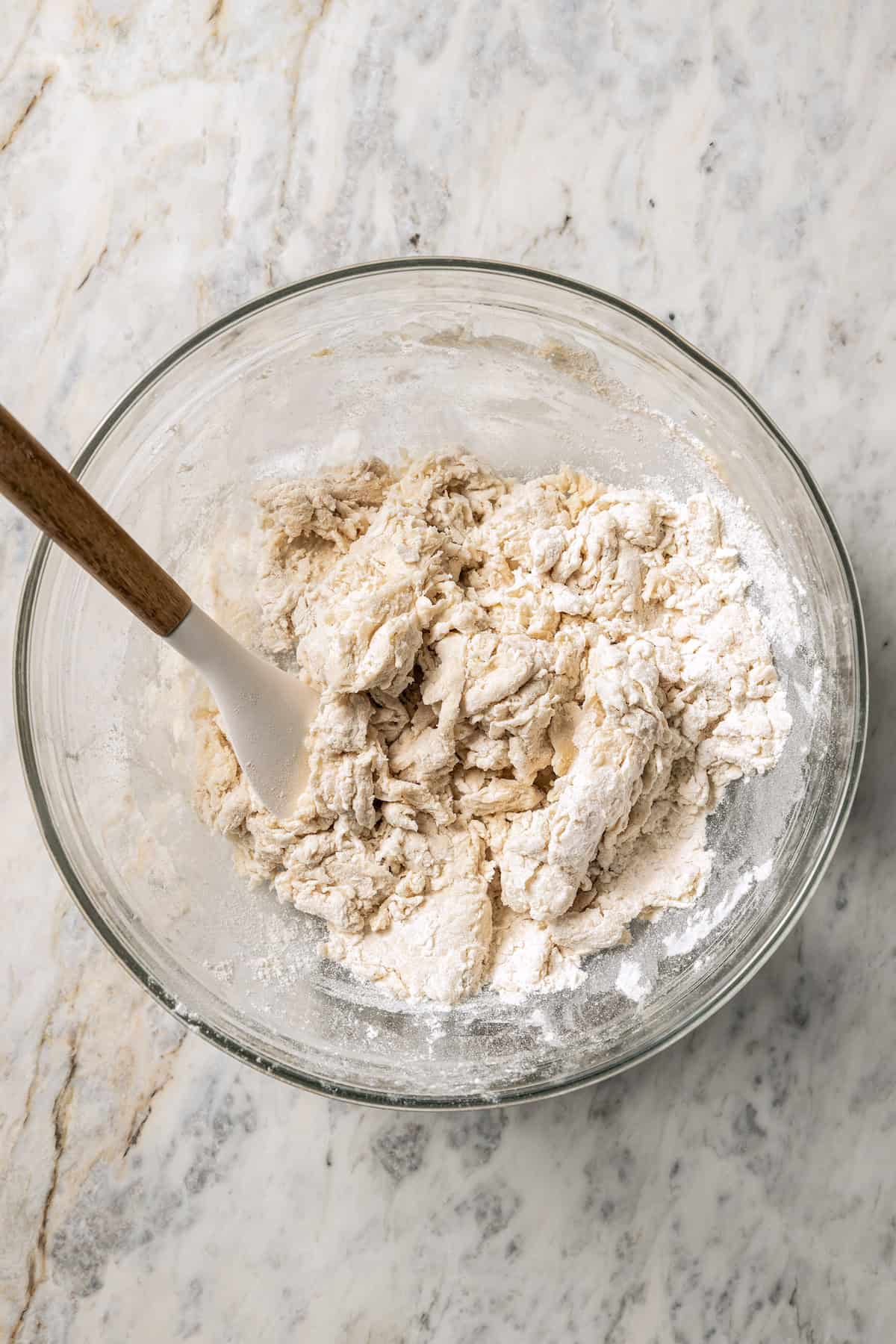 Shaggy dough in a glass bowl with a rubber spatula for stirring.