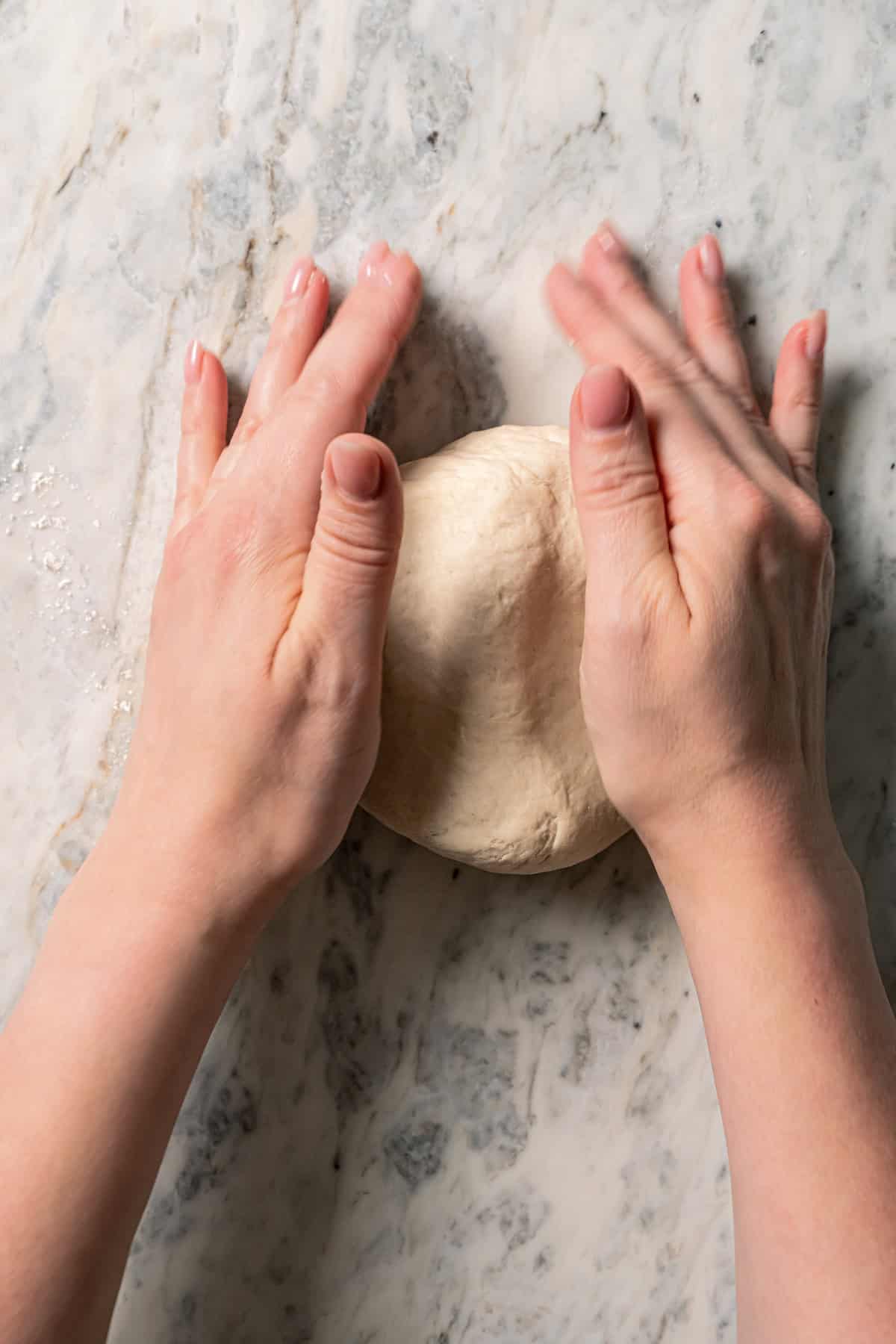 Two hands kneading bao bun dough on a marble countertop.