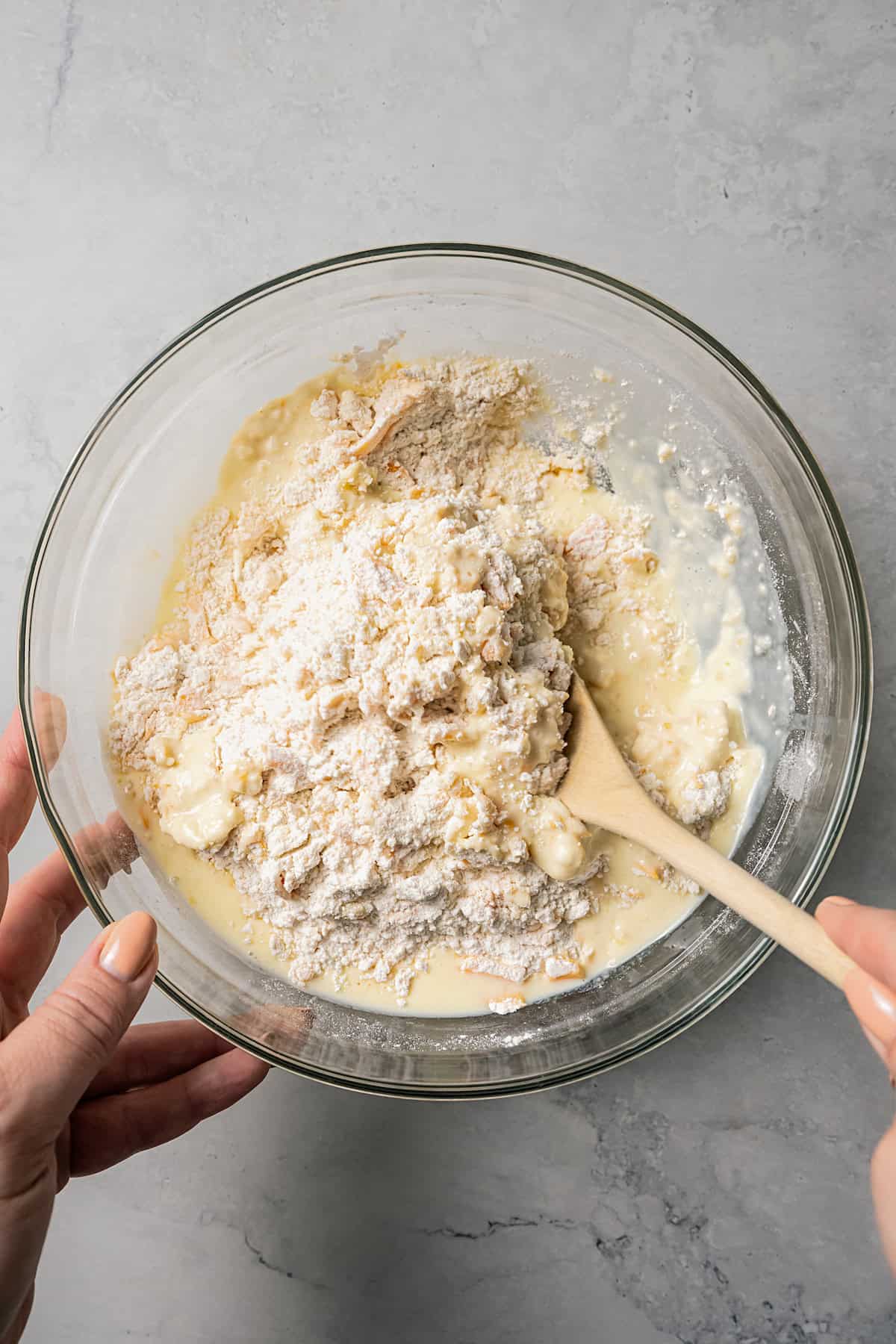 A hand holding a glass bowl of biscuit batter steady while the other hand uses a wooden spoon to combine the batter.