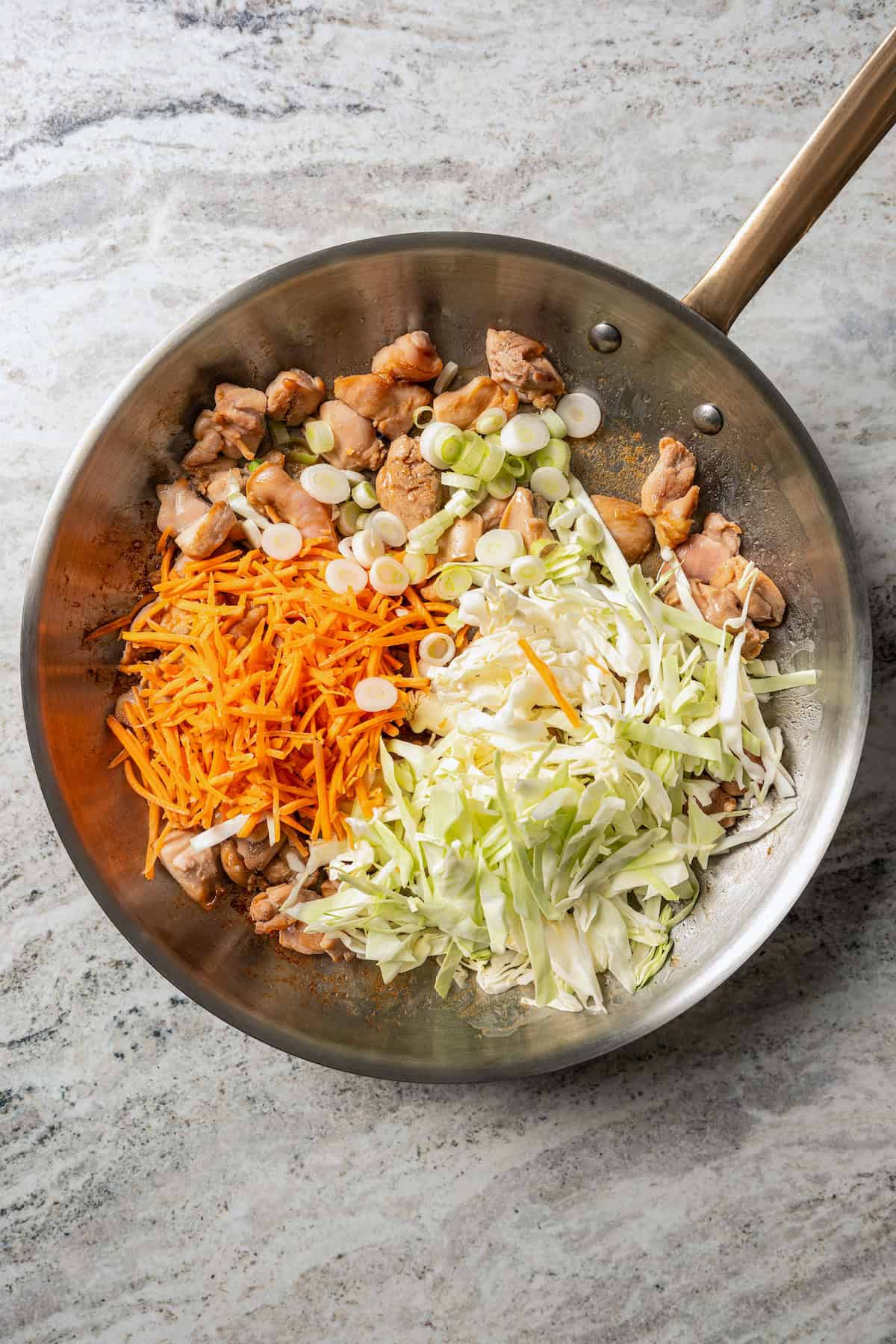 Shredded veggies added to chicken pieces in a stainless steel skillet.