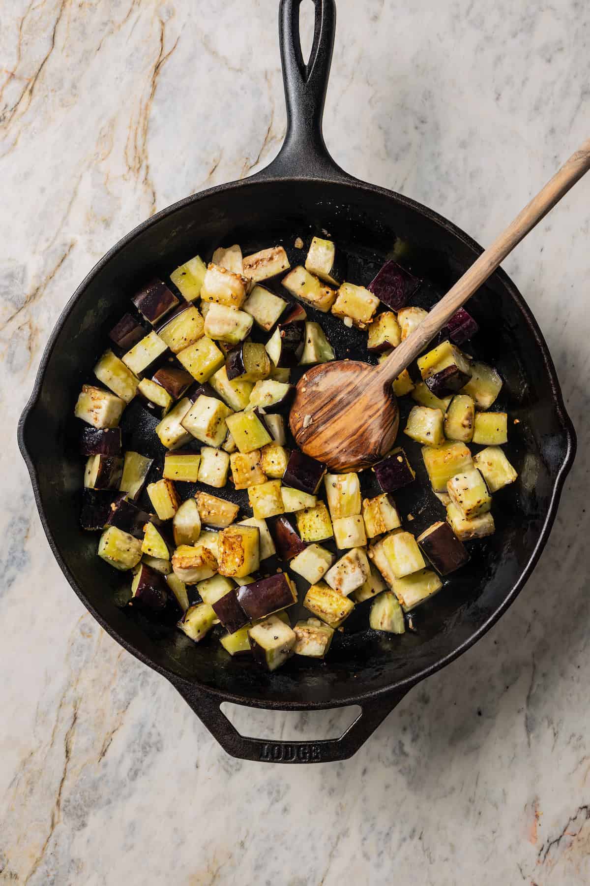 Sauteed eggplant cubes in a skillet with a wooden spoon.