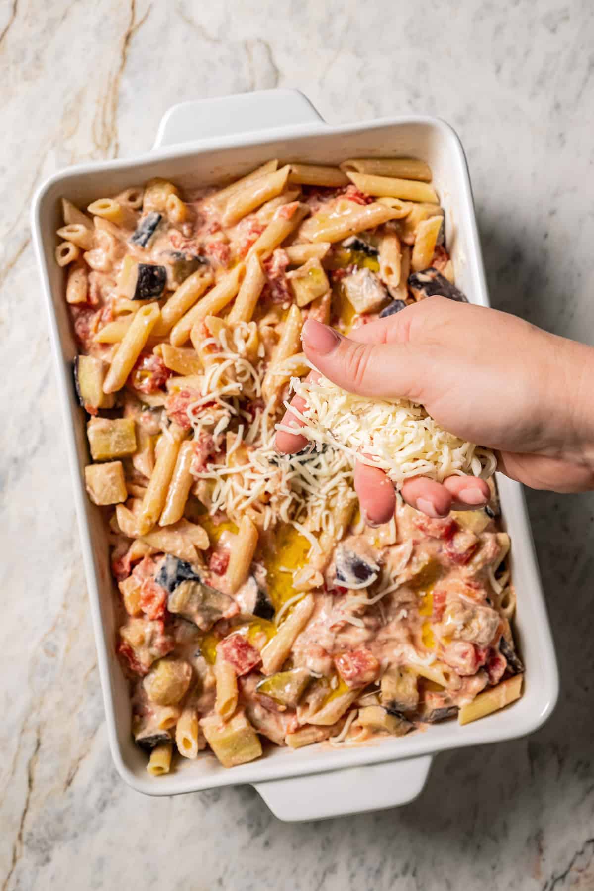 A hand sprinkling shredded mozzarella over eggplant penne pasta in a baking dish.