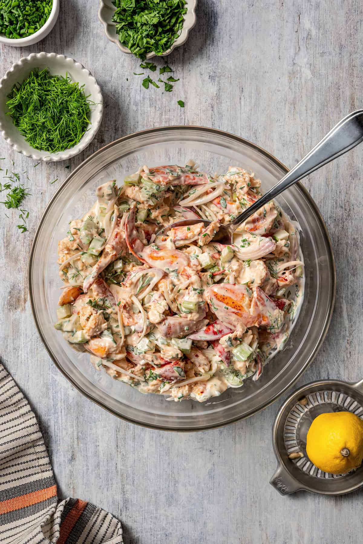 Lobster roll filling in a glass bowl with a stirring spoon, next to smaller bowls of chopped herbs and a lemon juicer.