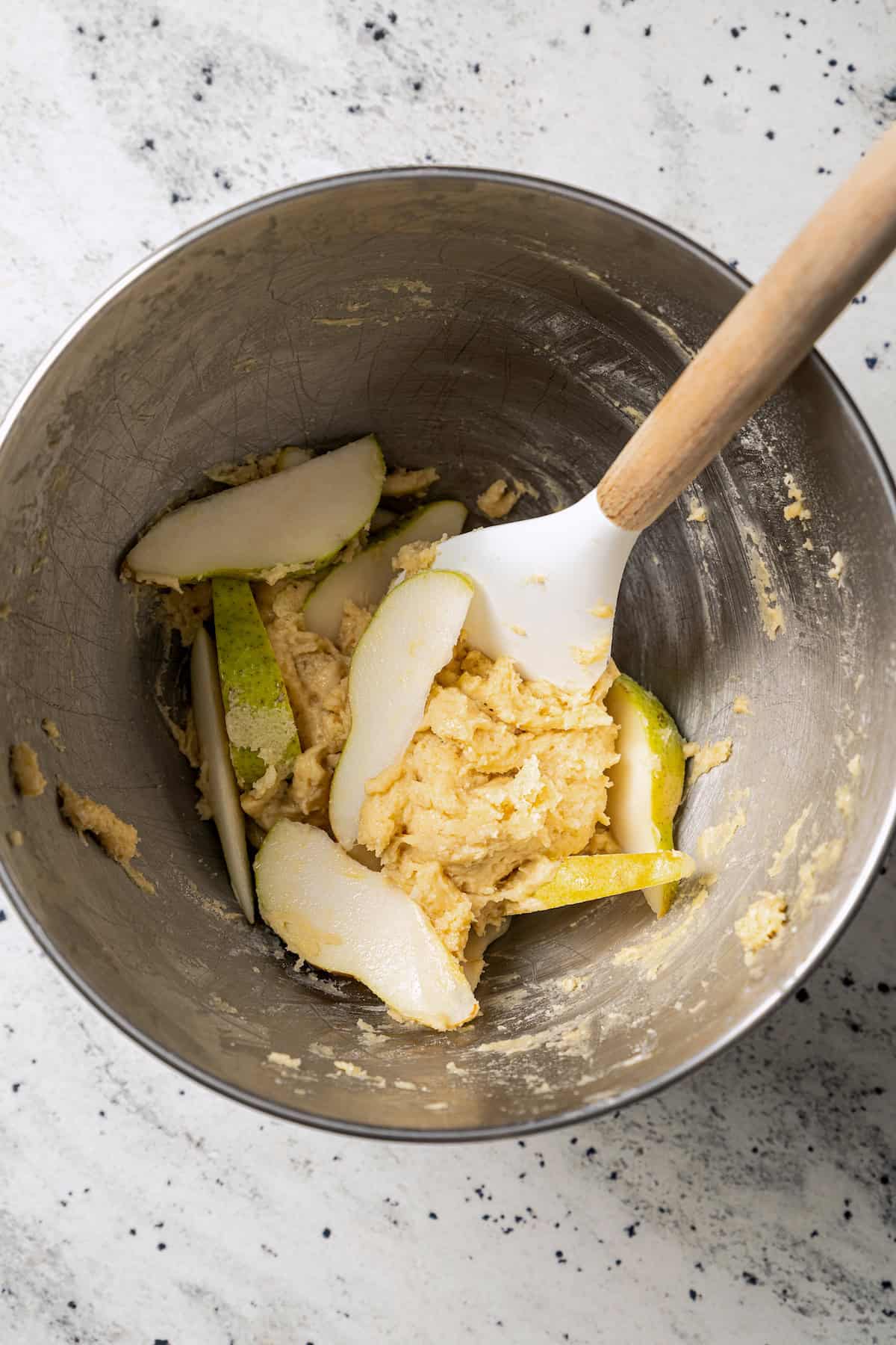 Sliced pears partially folded into a bowl of cake batter with a spatula.