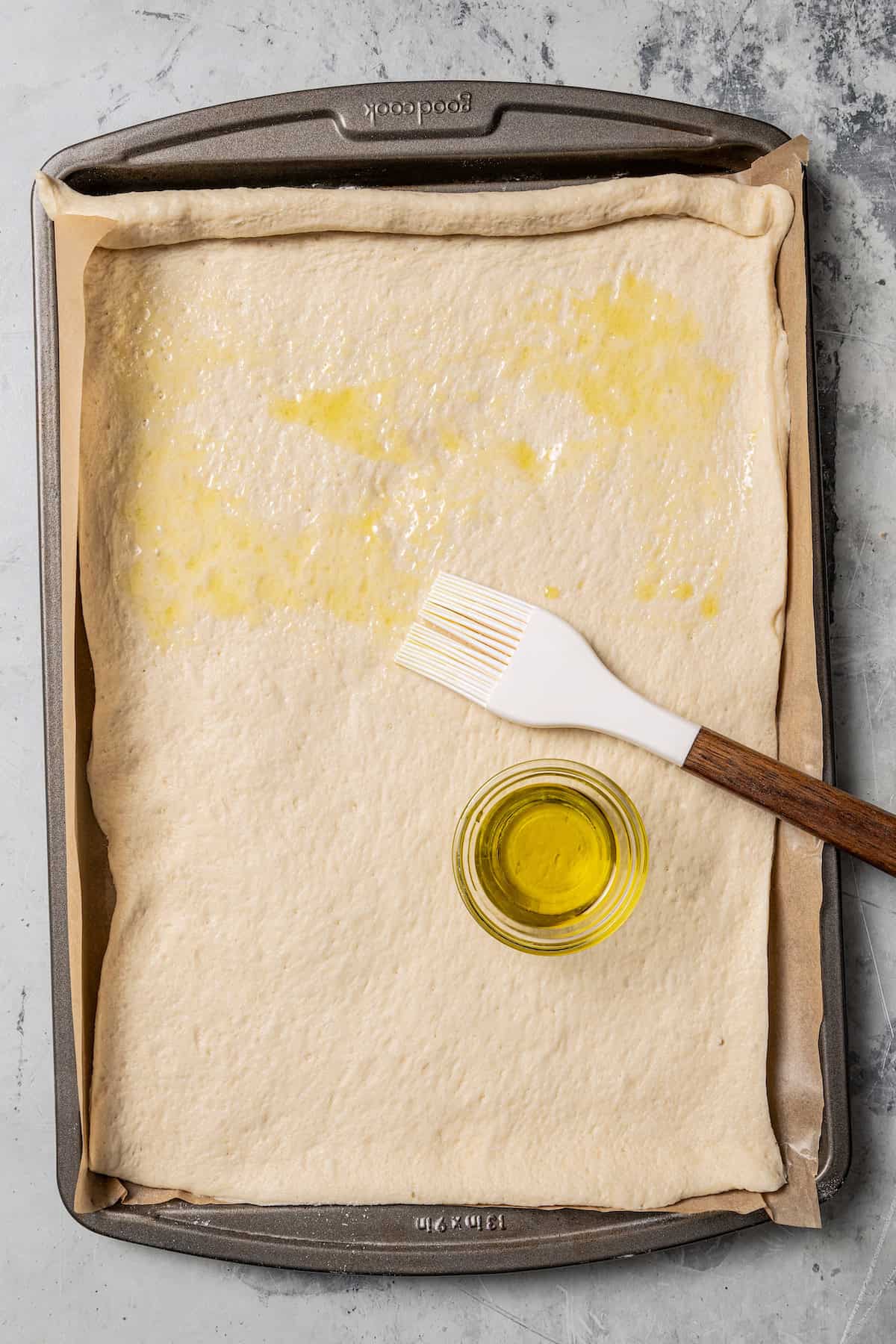 Overhead view of rolled out pizza dough being brushed with olive oil on a parchment-lined baking sheet.