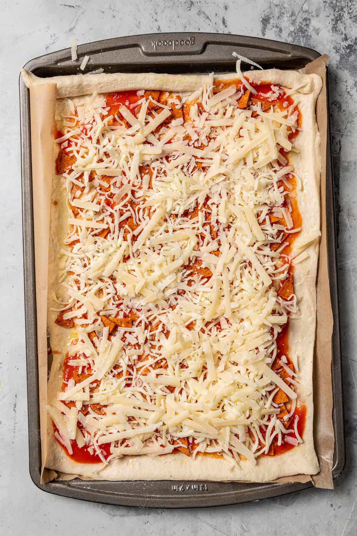 Overhead view of rolled out pizza dough topped with sauce and shredded cheese on a parchment-lined baking sheet.