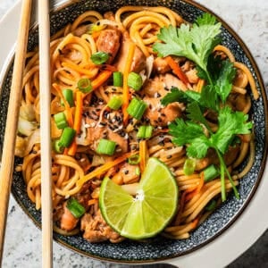 A bowl of chicken lo mein garnished with fresh herbs and lime wedges with a pair of chopsticks resting on the bowl.