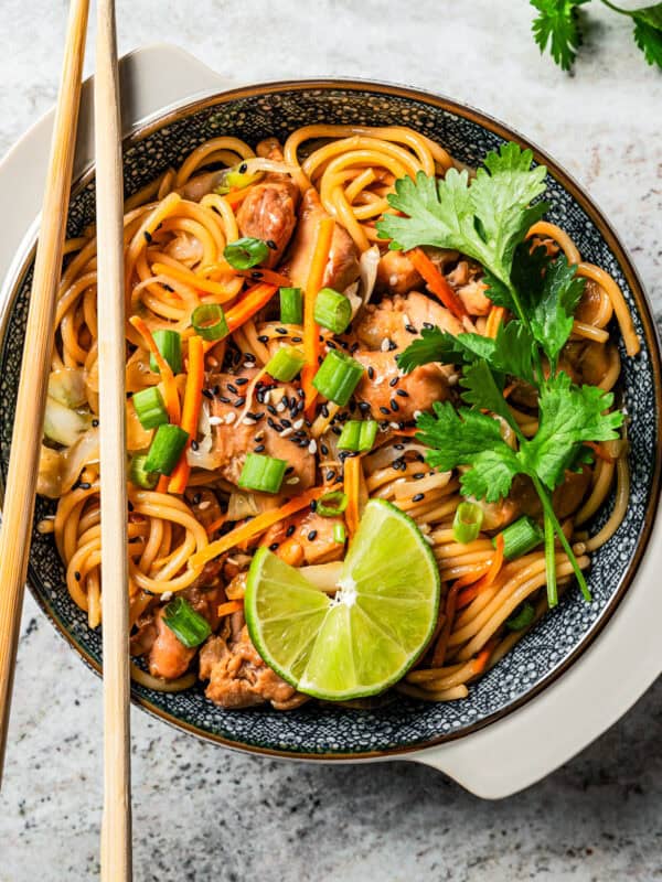 A bowl of chicken lo mein garnished with fresh herbs and lime wedges with a pair of chopsticks resting on the bowl.