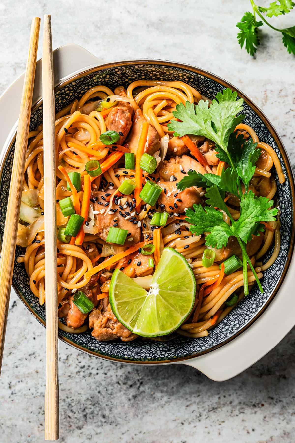 A bowl of chicken lo mein garnished with fresh herbs and lime wedges with a pair of chopsticks resting on the bowl.