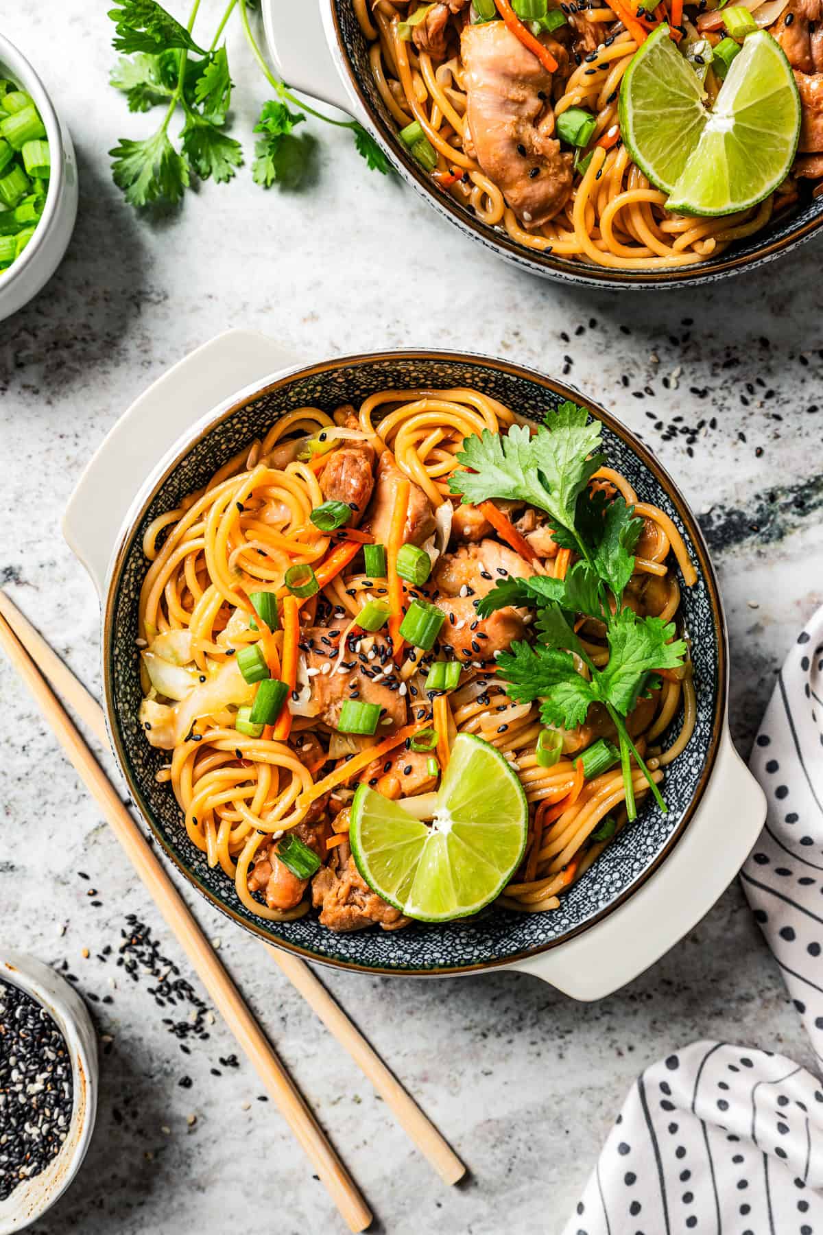 Overhead view of bowls of chicken lo mein garnished with fresh herbs and lime wedges, next to a pair of chopsticks and a second bowl of noodles.