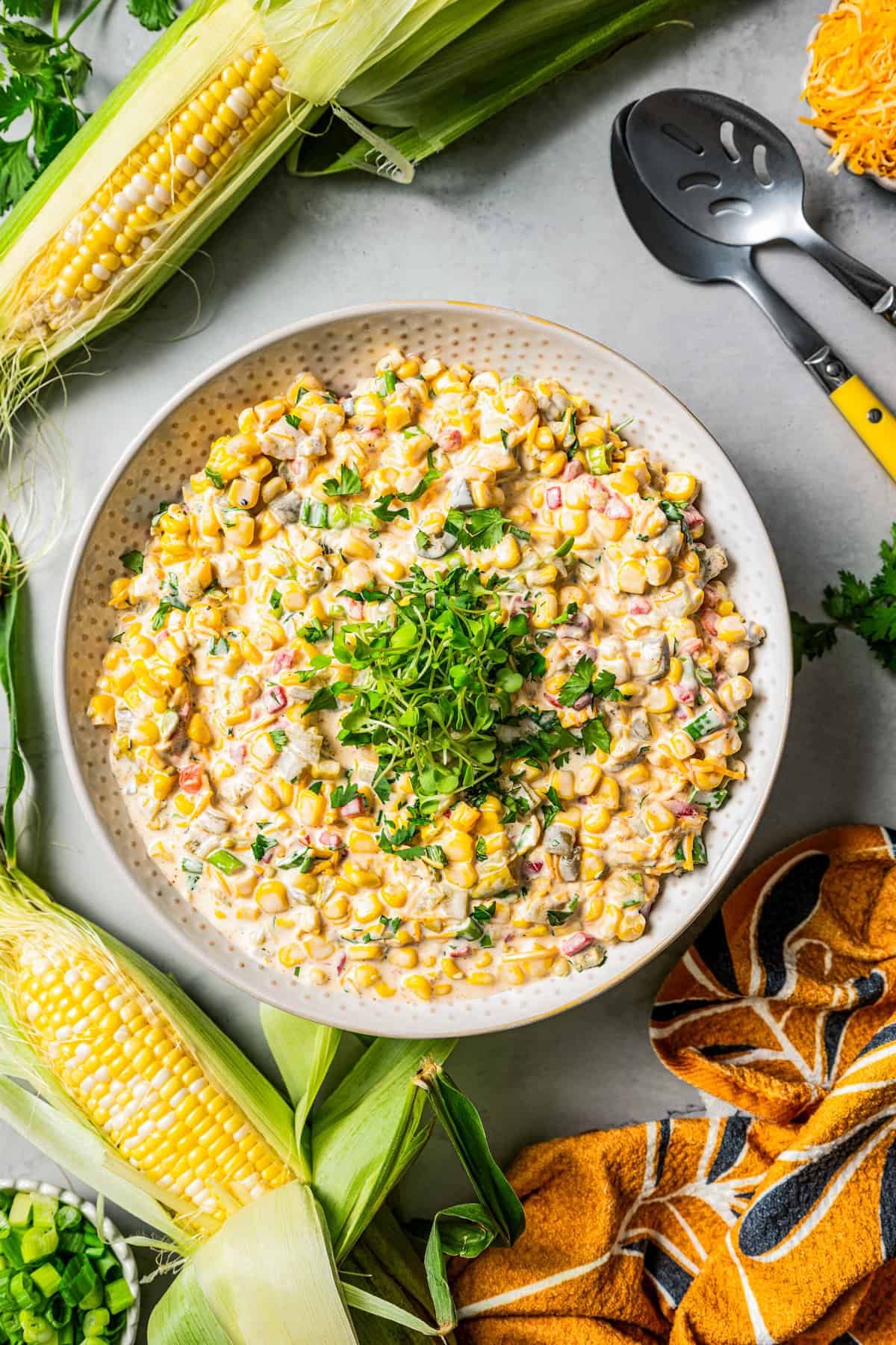 Overhead view of corn dip garnished with chopped herbs in a bowl, next to partially shucked ears of corn on the sob.