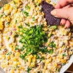 A hand dipping a black tortilla chip into a bowl of corn dip garnished with chopped herbs.