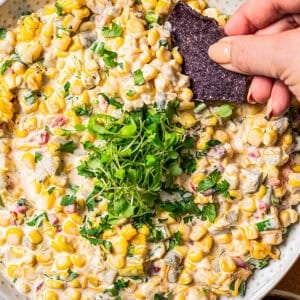 A hand dipping a black tortilla chip into a bowl of corn dip garnished with chopped herbs.