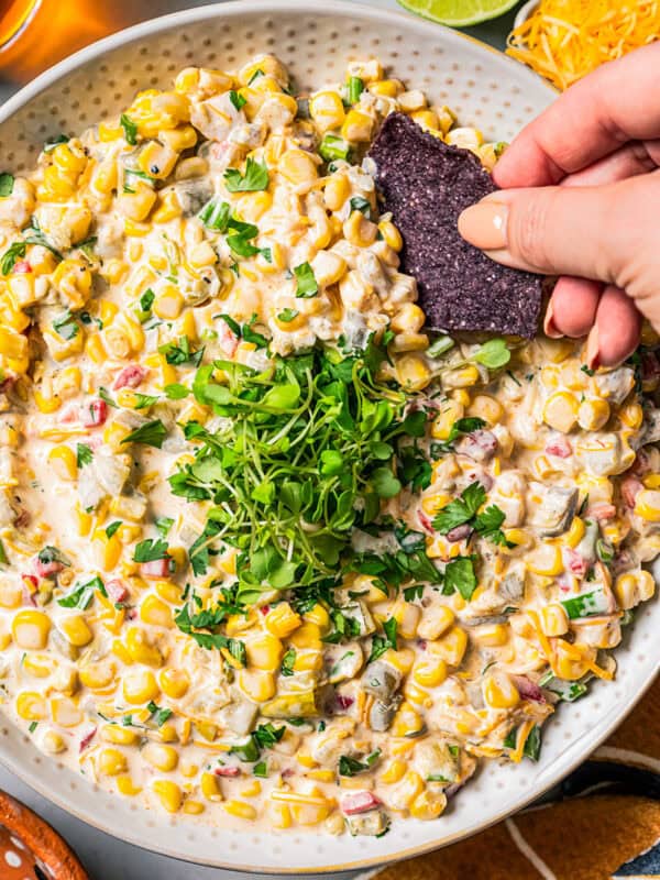 A hand dipping a black tortilla chip into a bowl of corn dip garnished with chopped herbs.