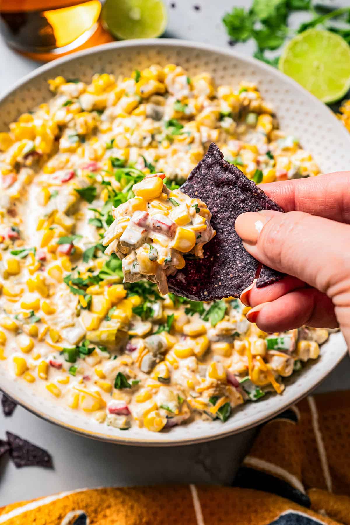 A hand holding up a black tortilla chip dipped in corn dip, with a bowl of dip in the background.