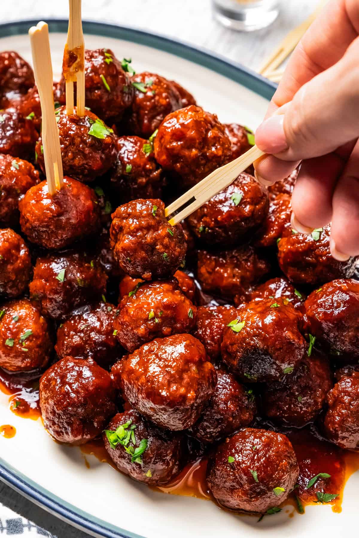 A hand using a cocktail skewer to pick up a grape jelly meatball from a large platter of meatballs.