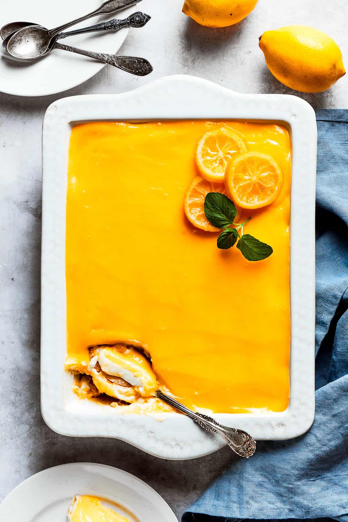 Lemon tiramisu in a white baking dish garnished with lemon slices and fresh green herbs, with lemons and dessert plates and spoons set all around the dish.