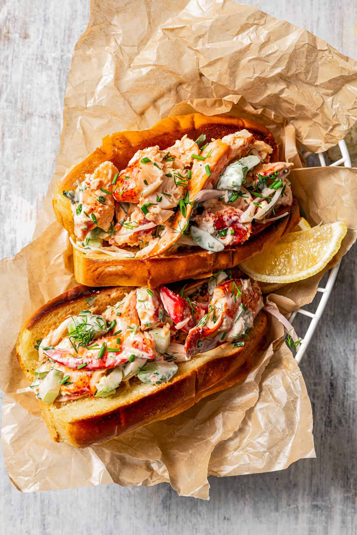 Overhead view of two homemade lobster rolls in a paper-lined food serving basket with a lemon wedge for garnish.