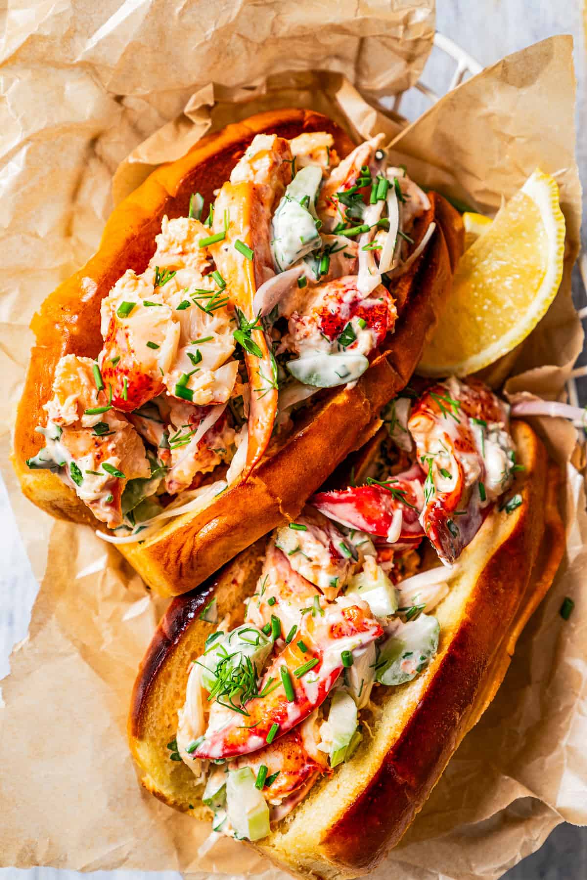 Overhead view of two homemade lobster rolls in a paper-lined food serving basket with a lemon wedge for garnish.