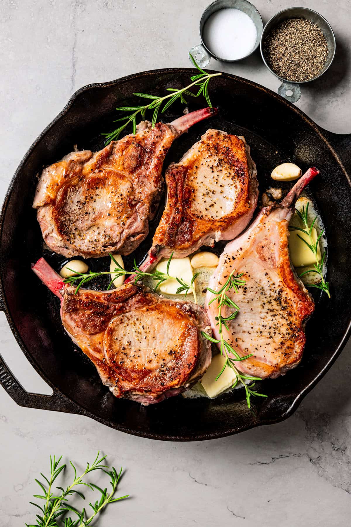 Herbs and butter added to a skillet with four seared tomahawk pork chops.