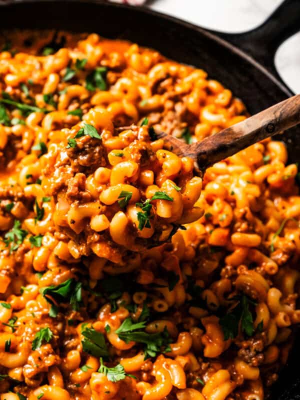 A serving spoon scooping macaroni pasta mixed with ground beef from a large skillet.