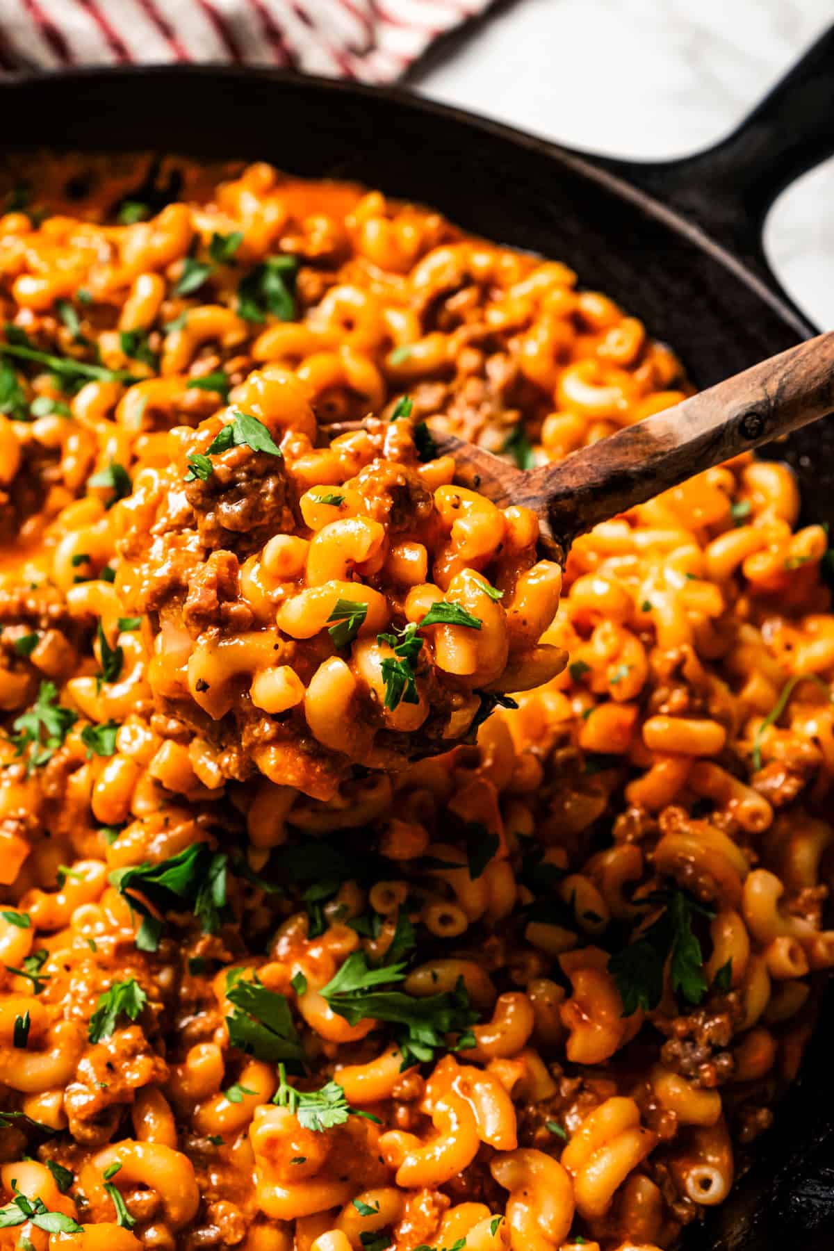 A serving spoon scooping macaroni pasta mixed with ground beef from a large skillet.