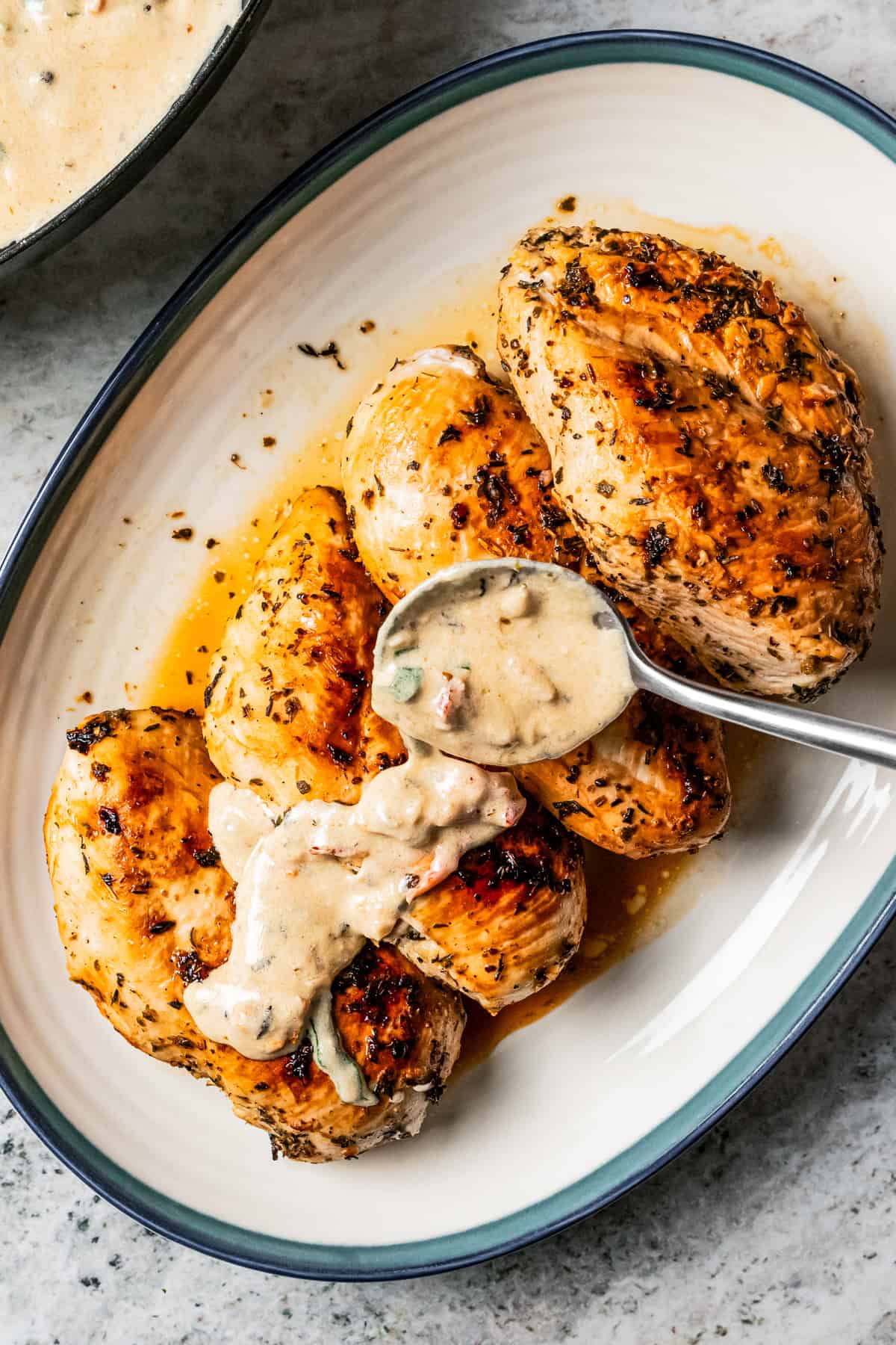 Lemon butter sauce is spooned over four cooked chicken breasts on a platter next to a skillet full of sauce.