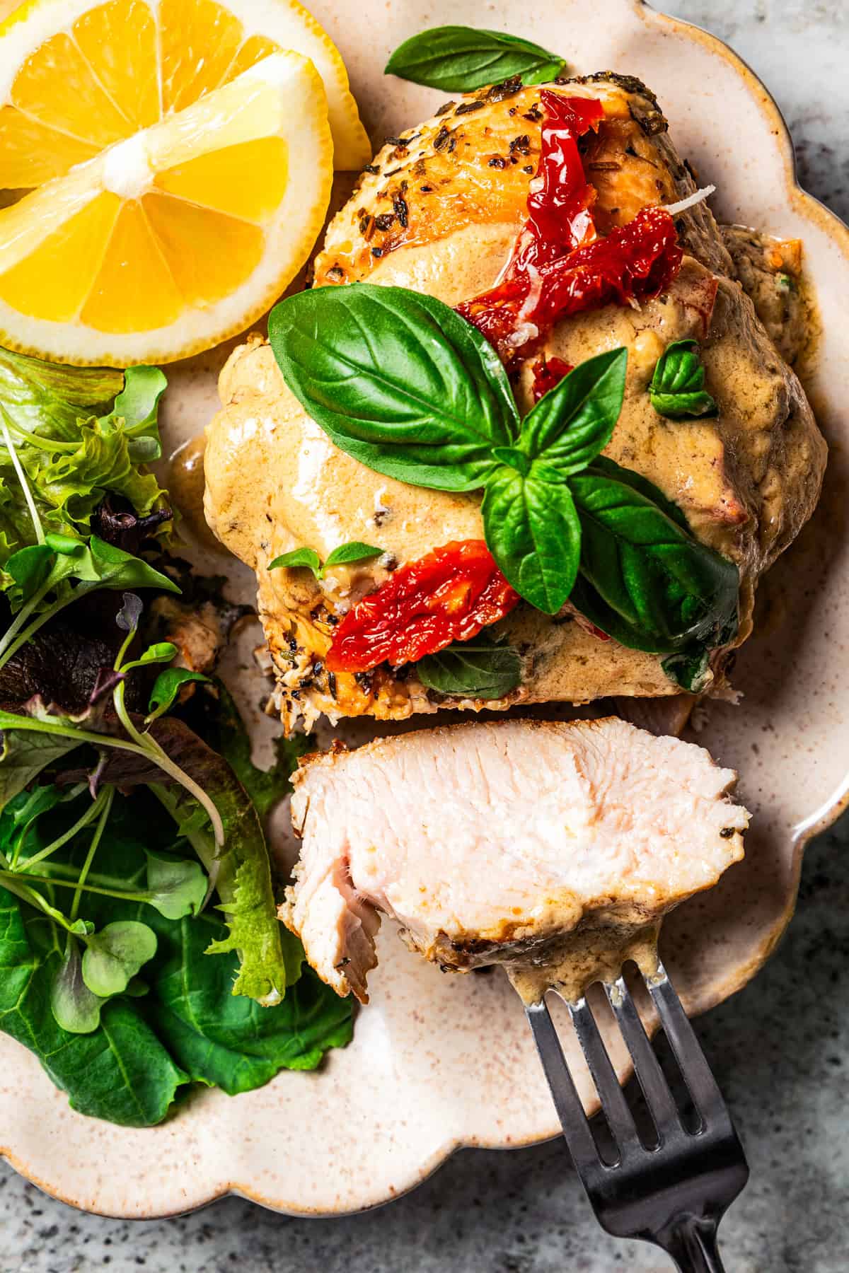 A fork pulls a slice of Chicken Bryan away from the rest of the chicken on a plate with a side of salad and lemon wedges.