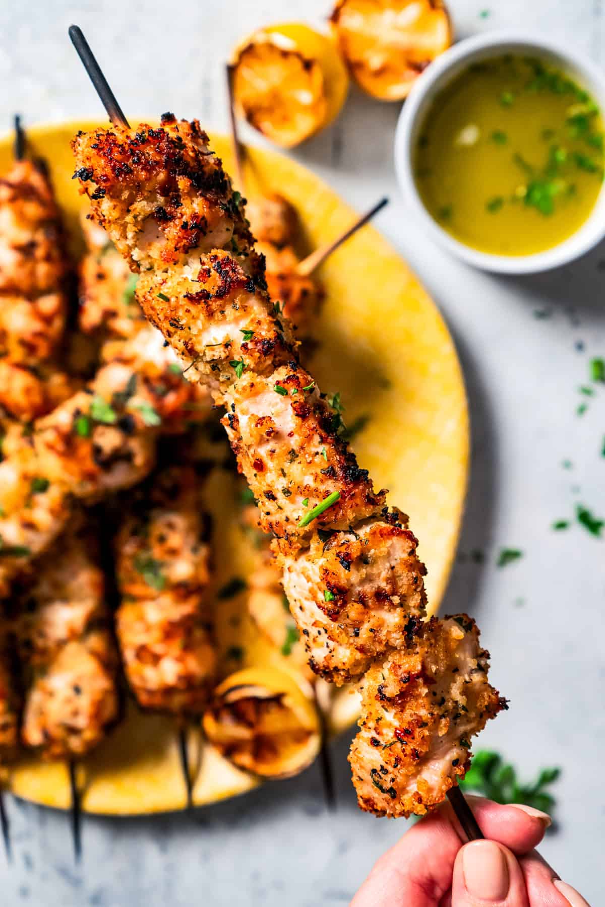 Close up of a hand holding up a chicken spiedini skewer, with more skewers on a yellow plate next to a small bowl of lemon butter sauce in the background.