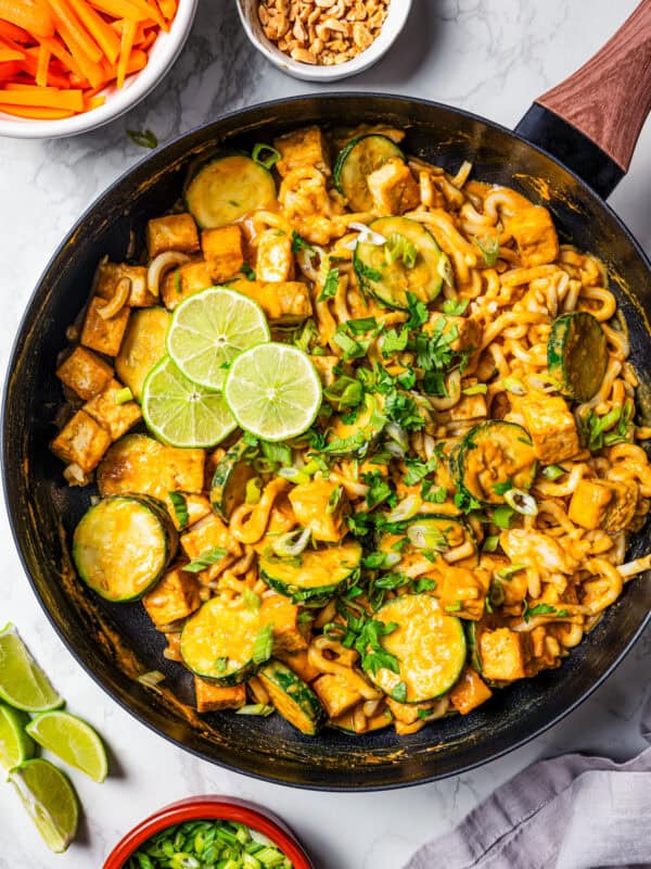 Peanut noodles with tofu in a dark skillet with bowls of peanuts, carrot sticks, and fresh herbs arranged around the skillet.