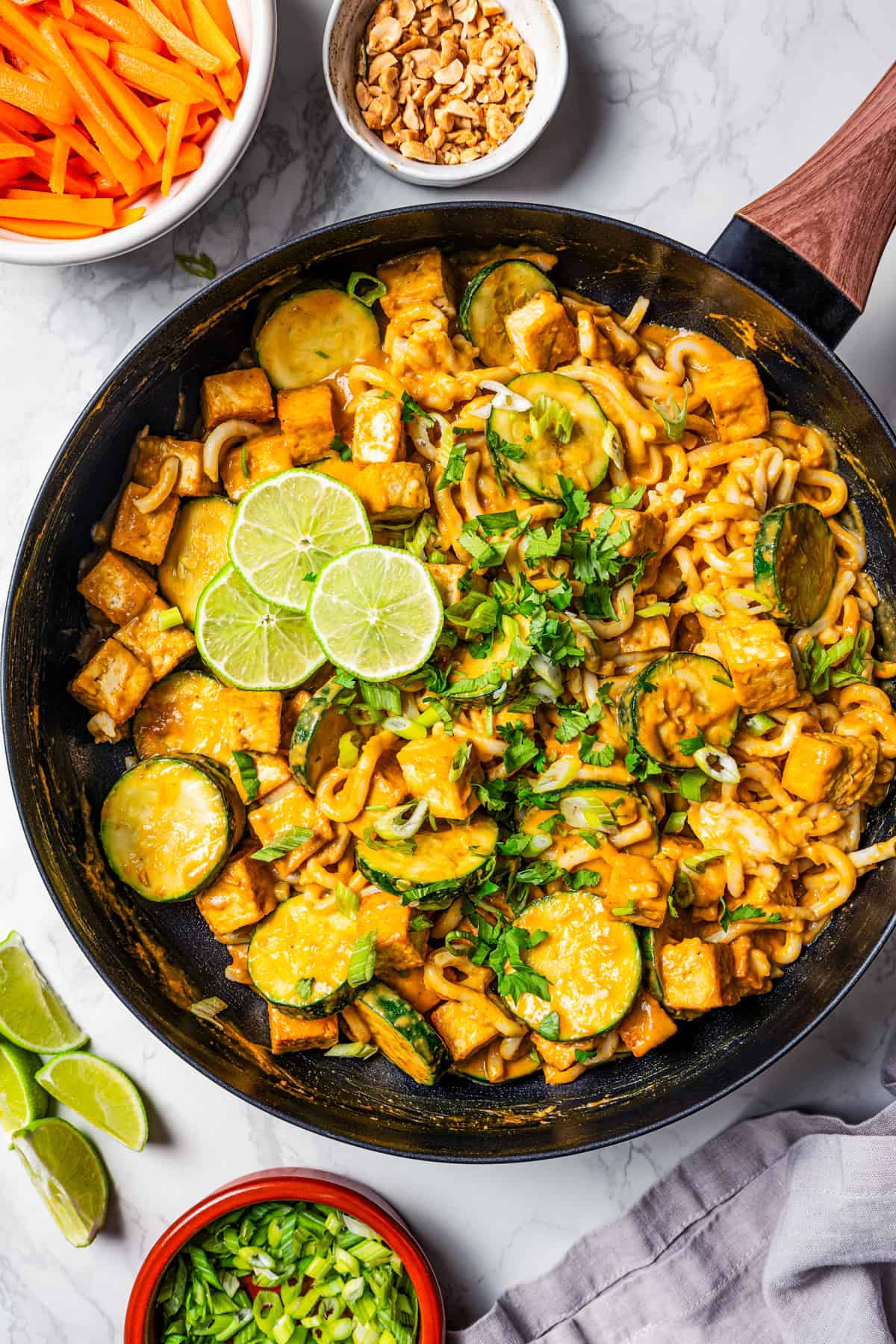 Peanut noodles with tofu in a dark skillet with bowls of peanuts, carrot sticks, and fresh herbs arranged around the skillet.