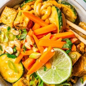 A bowl with peanut noodles tossed with cubed tofu, slices of zucchini, a garnish of carrot sticks, and a slice of lime.