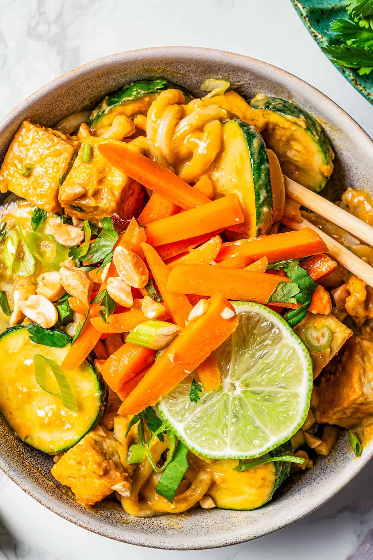 A bowl with peanut noodles tossed with cubed tofu, slices of zucchini, a garnish of carrot sticks, and a slice of lime.