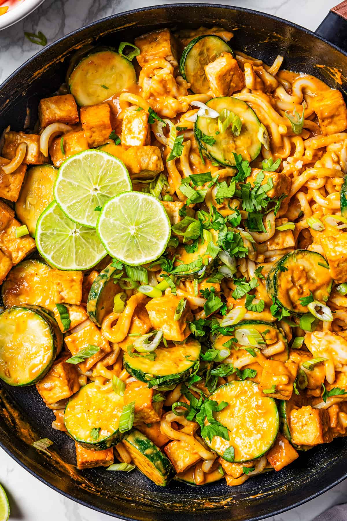 A pan of peanut noodles with tofu cubes and slices of zucchini.