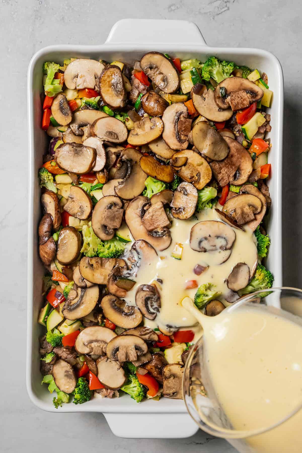 Egg mixture being poured over breakfast casserole ingredients assembled in a baking dish.