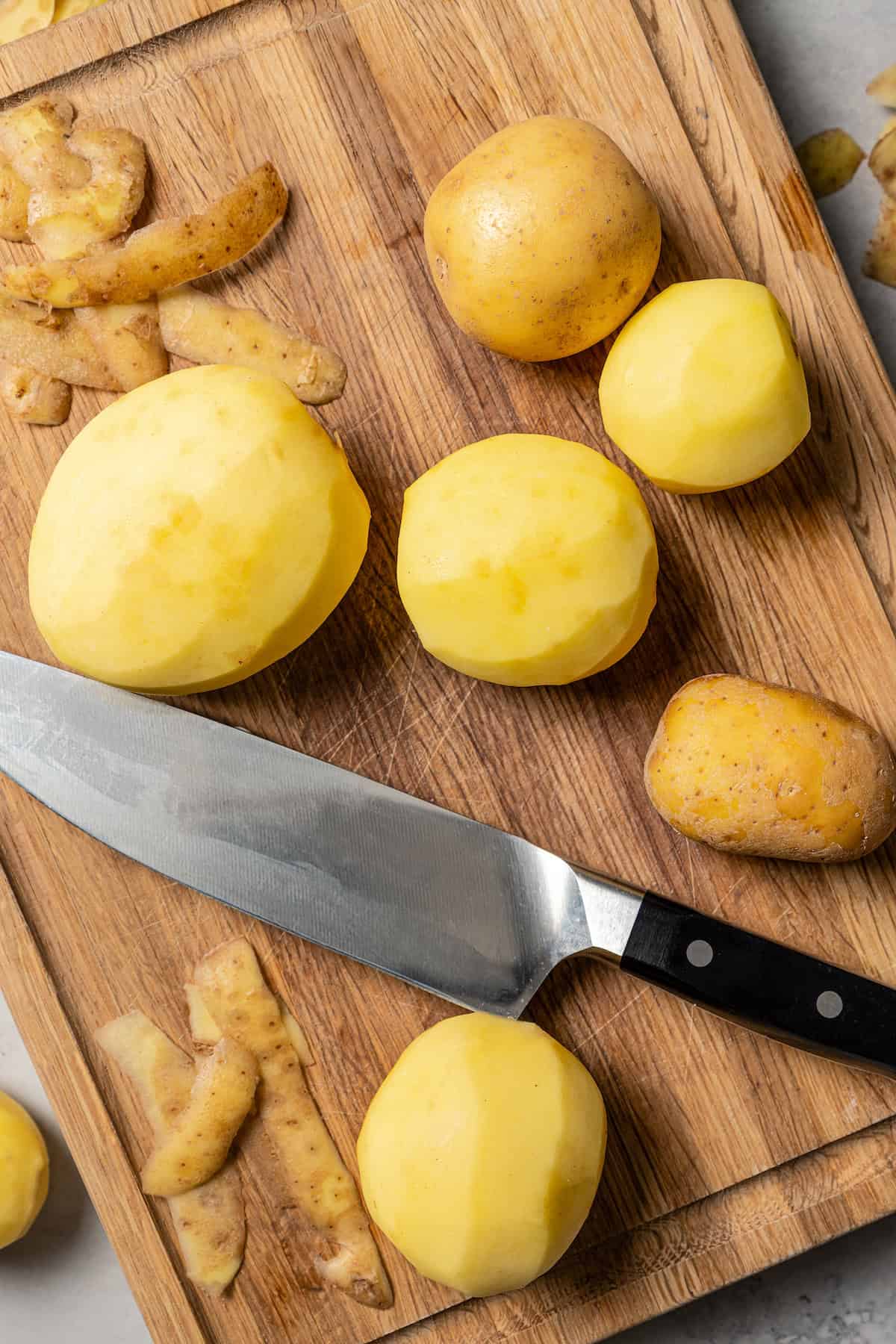 Peeled potatoes on a wooden cutting board next to a large knife.