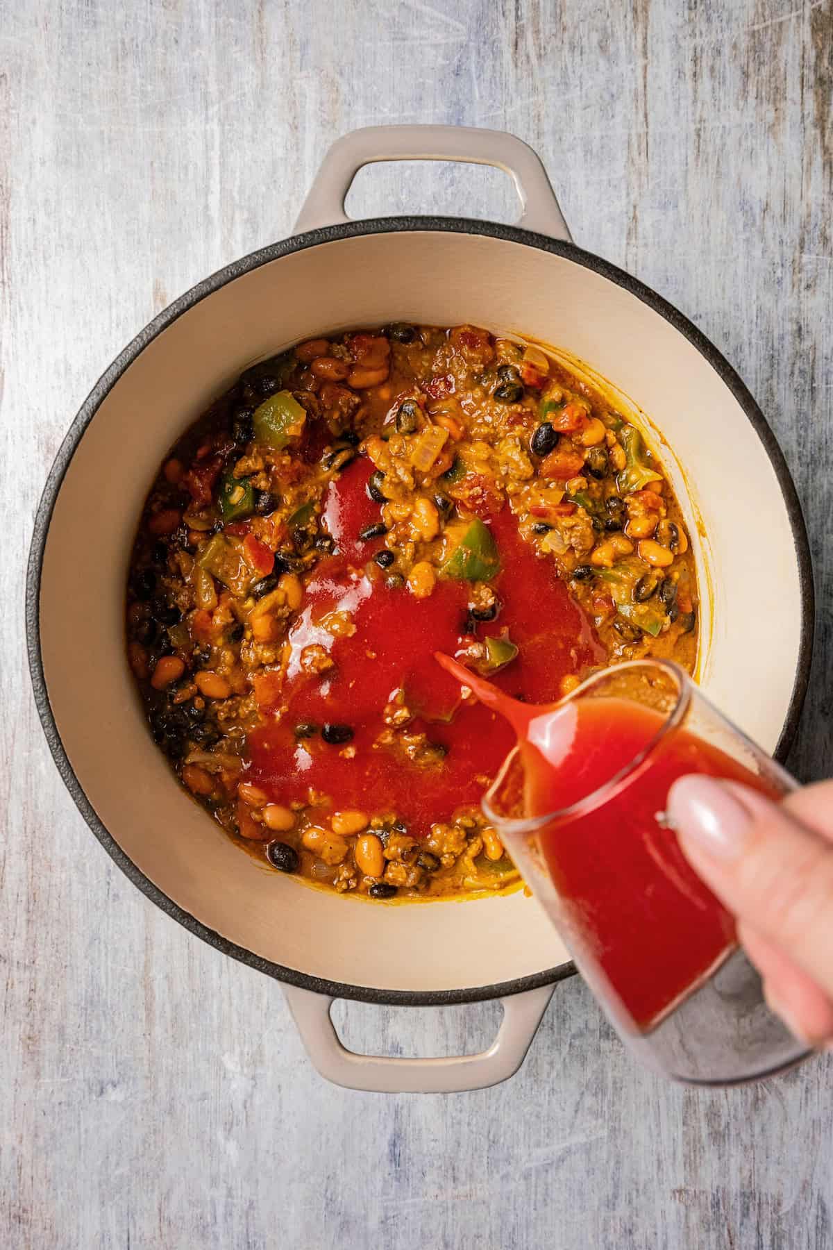 Tomato juice being poured into a large pot with pumpkin chili ingredients.