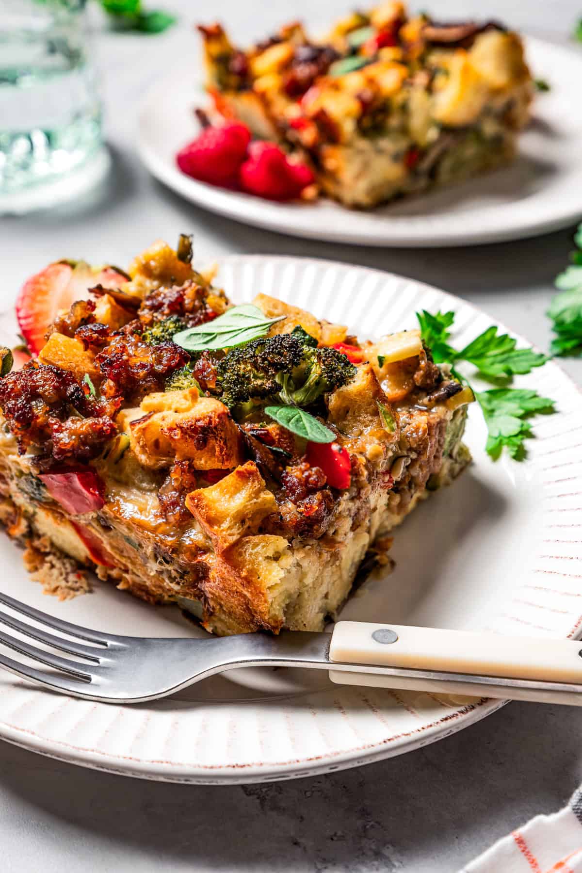 A slice of breakfast casserole with sausage on a plate next to strawberry halves and a fork, with a second slice on a plate in the background.