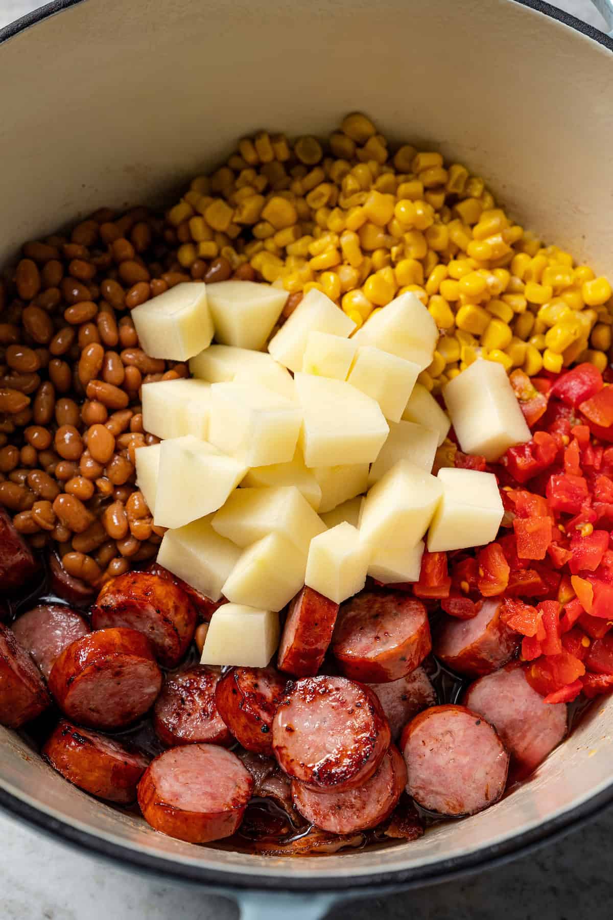 Kielbasa, bacon, ground beef, beans, corn, and potatoes combined in a soup pot.