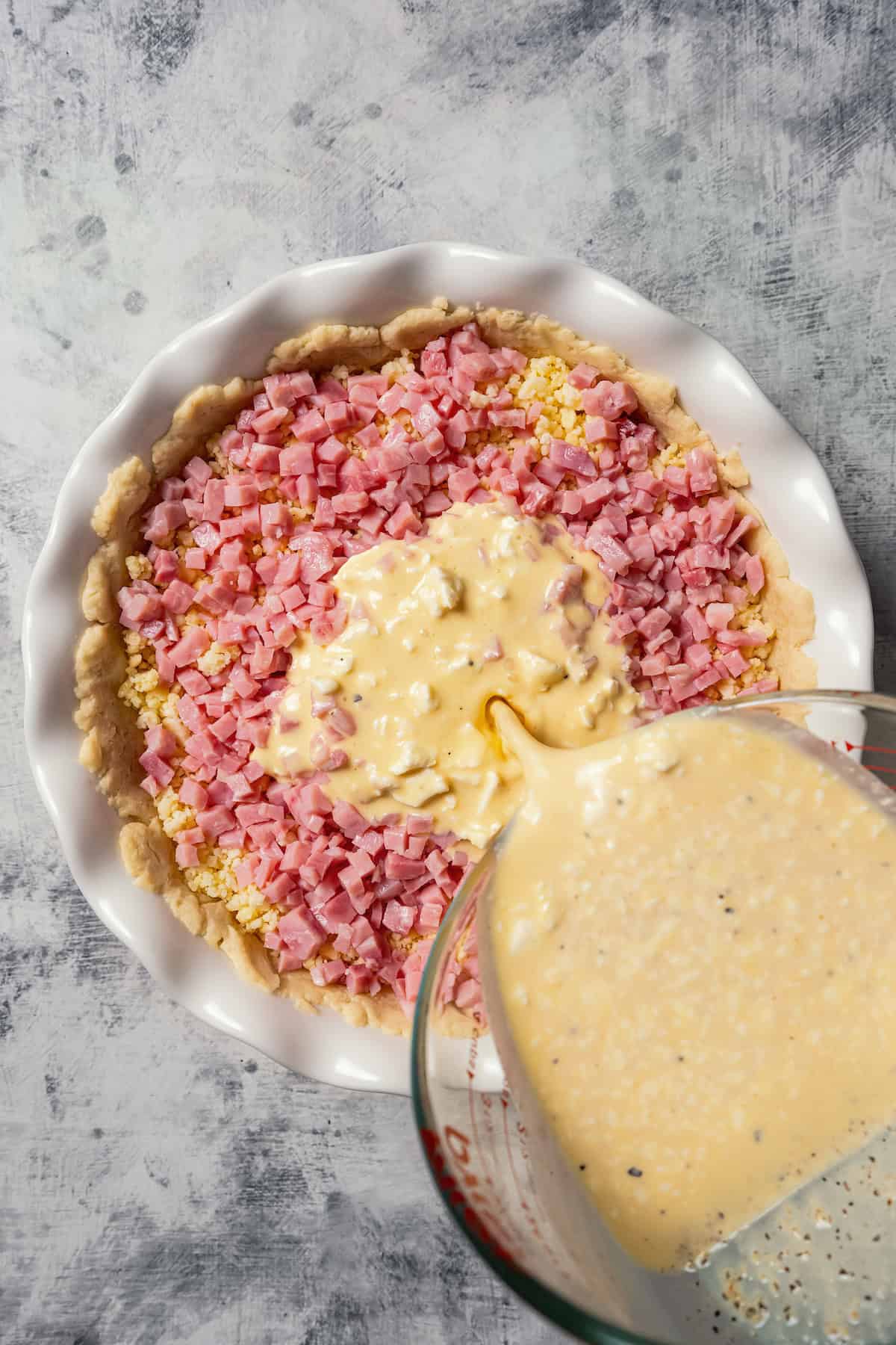 Quiche mixture being poured over ham and cheese in a pie crust.