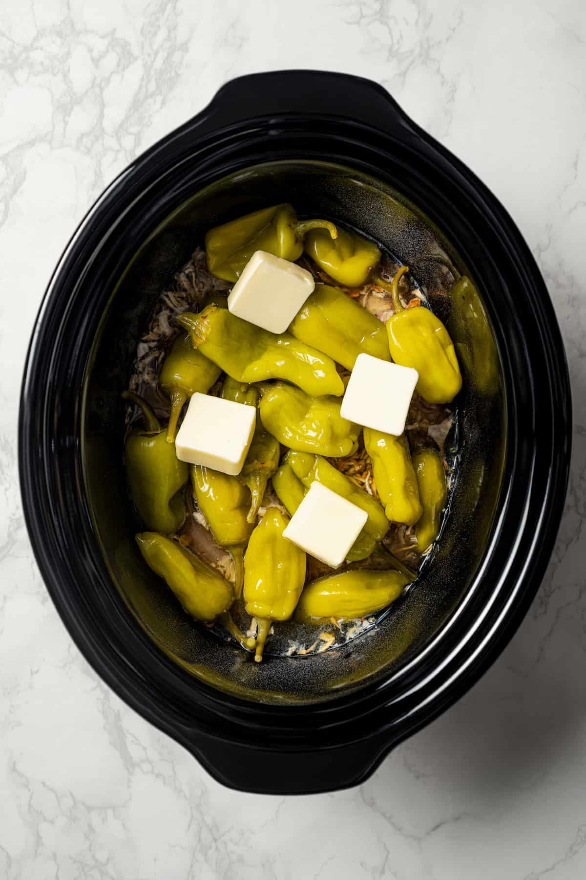 Pats of butter placed over top pepperoncini peppers and chicken inside the crock pot.