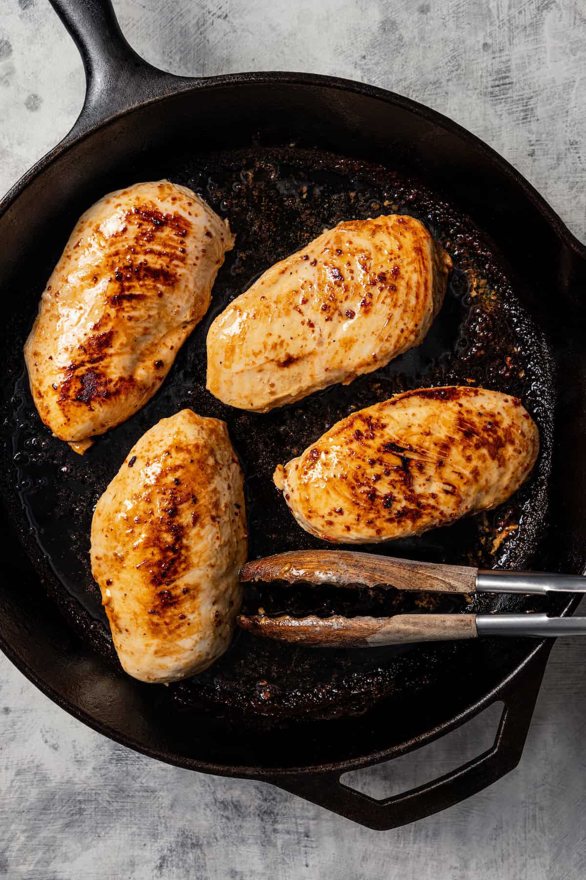 Browned chicken breasts in a cast iron skillet with a pair of tongs.