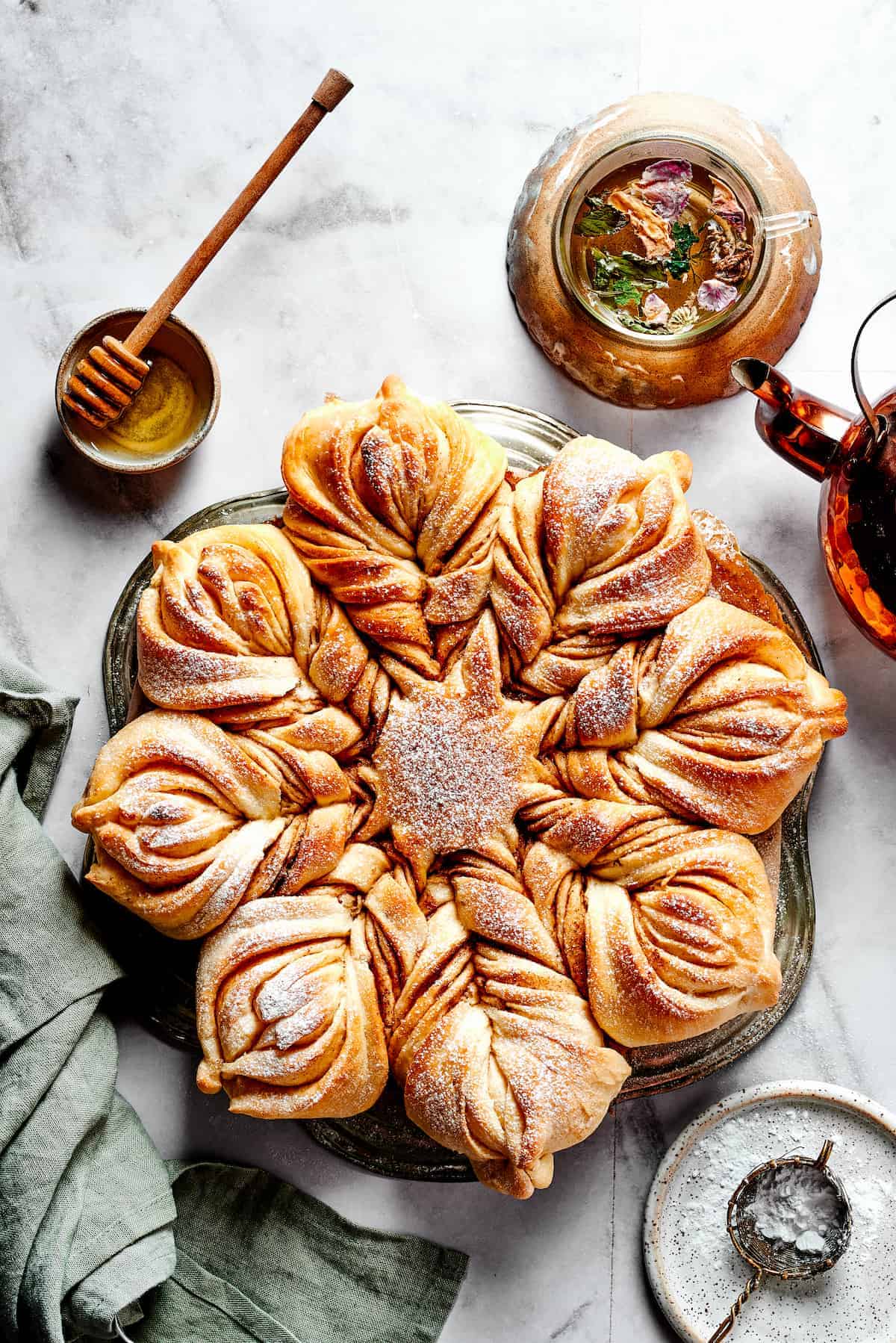 A bread shaped into a big star and served on a plate with a dusting of powdered sugar, and other ingredients set near the bread.