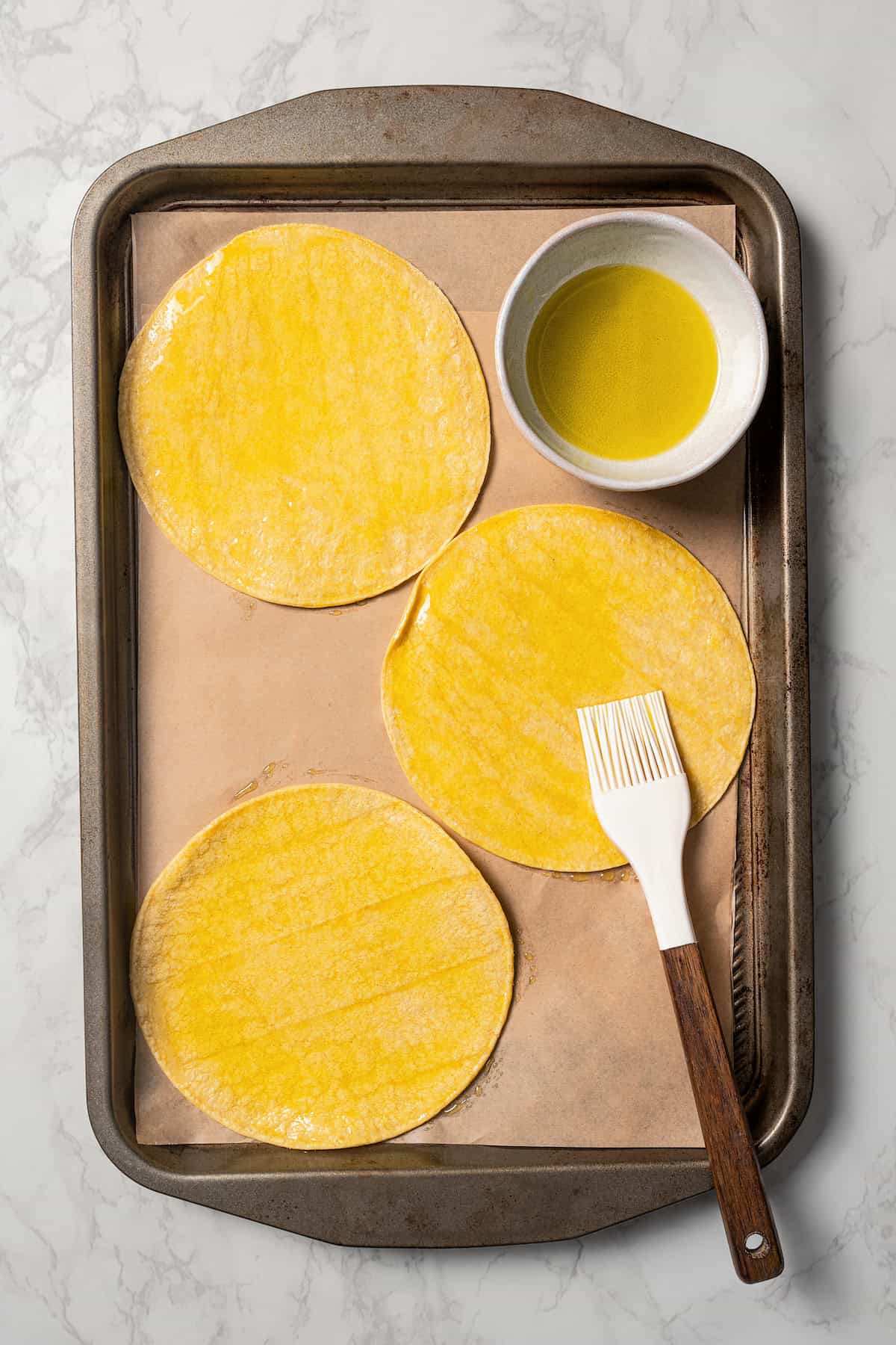 Overhead view of unbaked tostadas with a basting brush and a small bowl of oil on a lined baking sheet.