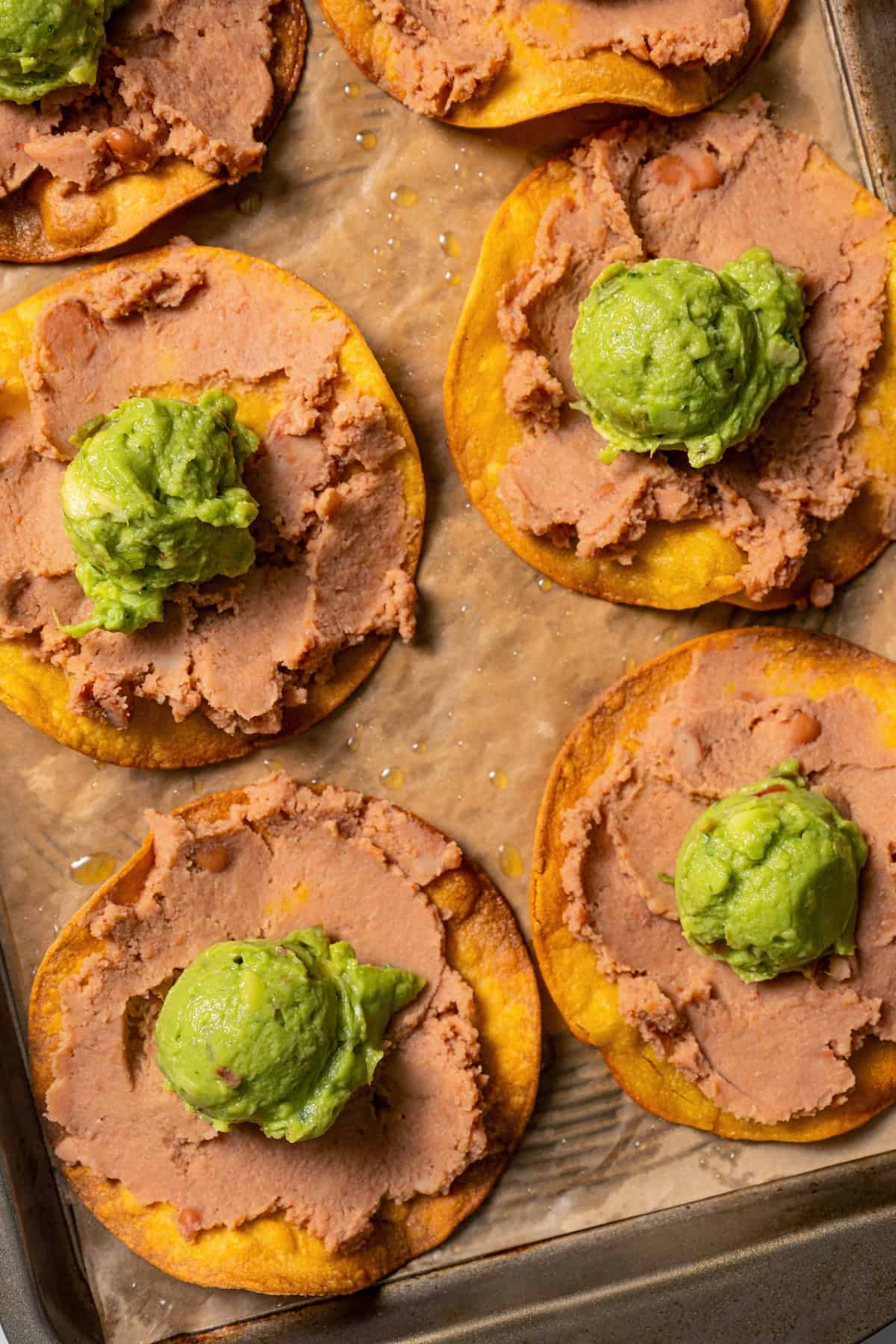 Overhead view of tostadas topped with refried beans and guacamole on a lined baking sheet.