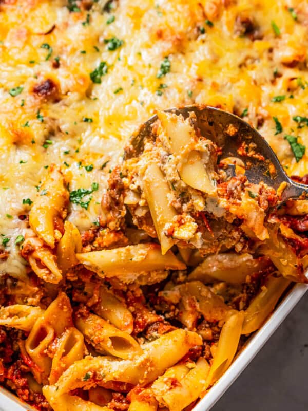 A spoon scooping baked mostaccioli pasta from the corner of a casserole dish.