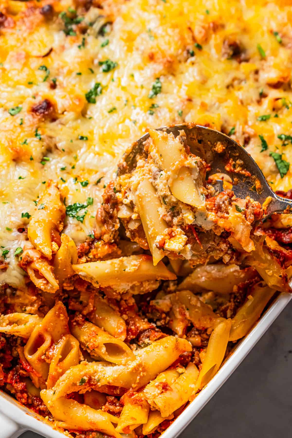 A spoon scooping baked mostaccioli pasta from the corner of a casserole dish.