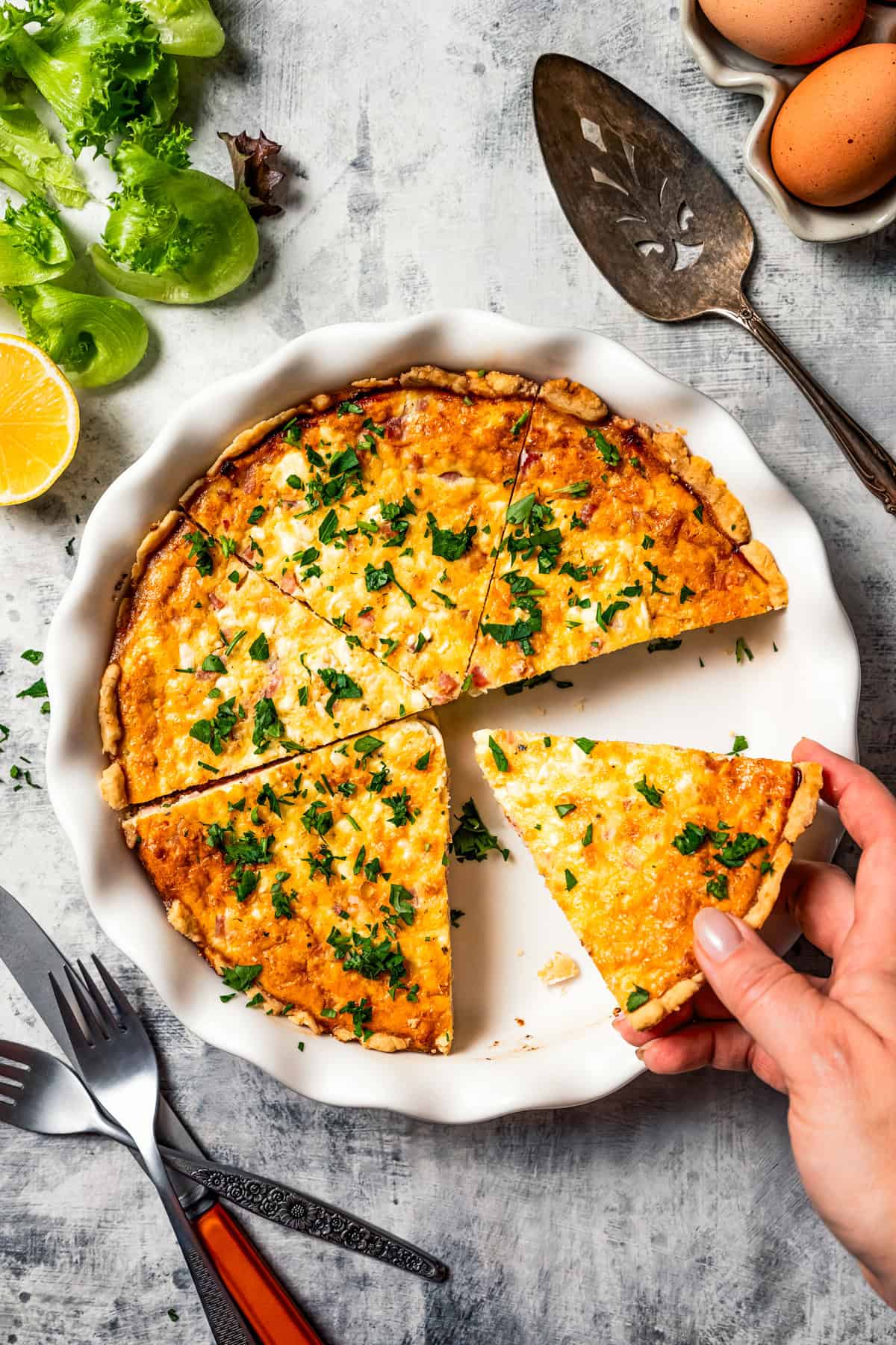 A hand lifting a slice of ham and cheese quiche from the rest of the quiche in a pie plate.